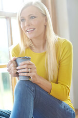 A beautiful woman smiling and sitting near a window inside a house. Happy attractive blonde woman chilling drinking coffee. Happy female relaxing and daydreaming at home off the grid in a sunroom