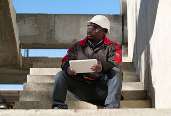 Tired african american worker sits at construction site and holds tablet PC