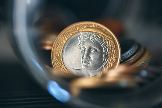 Real, Dinheiro do Brasil. Moedas Brasileiras dentro de um pote de vidro. Destaque para moeda de 1 Real. Economia Brasileira. Ambiente escuro. Fotografia macro.