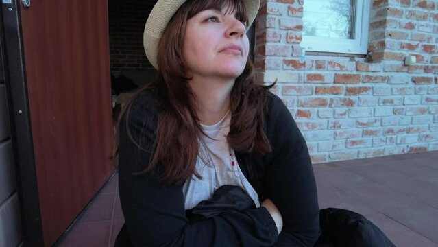 a woman in a hat sits on the porch of the house and thinks about something