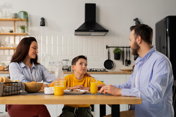 A little boy eats breakfast with his parents at the kitchen table. A boy in a yellow jacket communicates with his parents during breakfast.