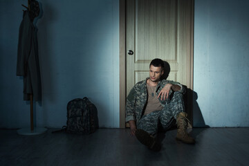 Frustrated military veteran in uniform sitting on floor near door in hallway at night.