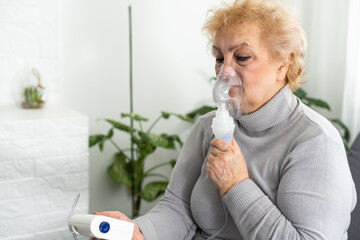 Sick senior woman has a viral infection or flu, labored breathing, asthma. An elderly female is using inhaler, nebulizer for self treatment sitting alone on the sofa at home