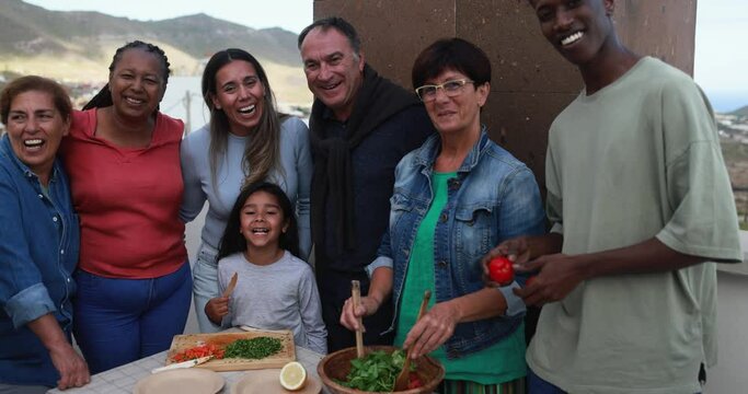 Multiracial friends having fun smiling in front of camera while preparing food with child during weekend day - Multi generational people doing barbecue at home's rooftop while smiling on camera