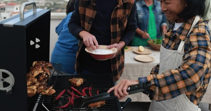 Multi generational people doing barbecue during weekend day at home's rooftop preparing food - Multiracial friends having fun together - Mature asian woman cooking chili on bbq grill
