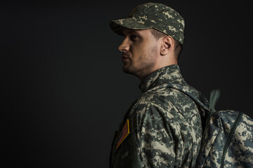 patriotic soldier in camouflage uniform standing with backpack isolated on black.
