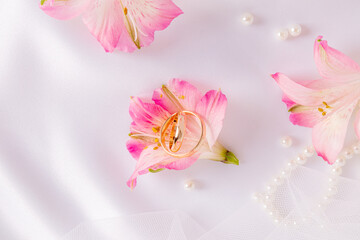 Two gold engagement rings inside a lovely pink astromeria flower on a white satin background with pearls. Wedding background. A copy space.