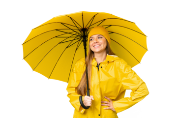Young blonde woman with rainproof coat and umbrella over isolated chroma key background posing with arms at hip and smiling