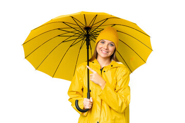 Young blonde woman with rainproof coat and umbrella over isolated chroma key background pointing to the side to present a product © luismolinero