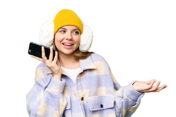 Young blonde woman wearing winter muffs over isolated chroma key background keeping a conversation with the mobile phone with someone