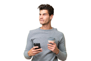 Young handsome caucasian man over isolated background holding coffee to take away and a mobile while thinking something