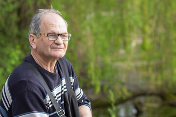 Senior man with glasses relaxing and breathing fresh air in spring park, green nature background. Walking and dreaming.