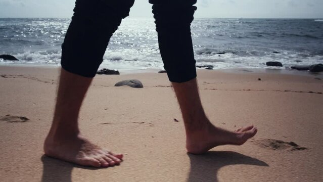 Close-up of bare male feet walking along the sandy coast on a summer day. Legs of a walking man on a sandy beach against the background of the sea, slow motion