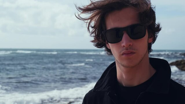 Young man with blown brown hair against the ocean on summer sunny day. Attractive young man looks at the camera on the beach. Handsome guy in black sunglasses poses outdoors on vacation. Slow motion