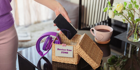 Close up of hand holding smartphone in wicker basket with inscription Device free zone. Woman putting phone into box with different gadgets at home. Digital detox and technology dependance concept