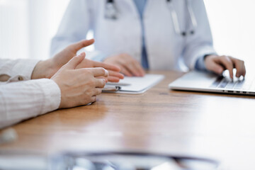 Doctor and patient sitting at the wooden desk in clinic. Female physician's hands pointing into laptop computer monitor, close up. Medicine concept