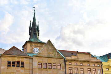 Old building with a green tower with a spire and a clock, ancient buildungs with yellow walls and green roofs on the foreground. Sunny day with blue sky. Prague, Czech Republic, October 2022.