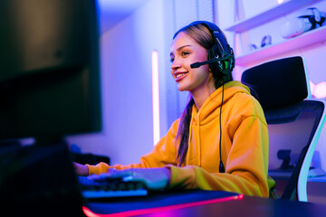 Young Caucasian woman professional gamer wearing yellow hoodie sits on a chair with a gaming table with pc, keyboard, monitor, microphone. Prepare for competition, cast gameplay or record a podcast.