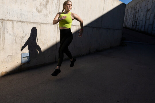 Fit sportswoman jogging on asphalt road by concrete wall