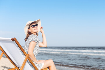 Happy brunette woman wearing sunglasses and hat relaxing on a wooden deck chair at the ocean beach