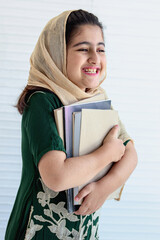 Portrait of happy smiling Pakistani Muslim student girl child with beautiful eyes wears hijab traditional costume, holding book on white background, kid female education, achieving gender equality.