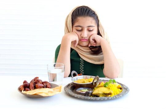 Adorable Pakistani Muslim girl with beautiful eyes sitting at kitchen table, kid wearing hijab and traditional costume feeling bored, Anorexia, with traditional Islamic halal food on white background.