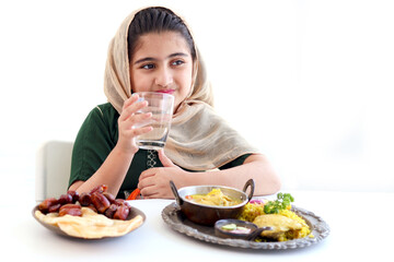 Adorable Pakistani Muslim girl sitting at kitchen table with traditional Islamic halal food, kid with hijab drinks water, enjoy eating traditional Islamic halal food with hand on white background.