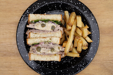 Closeup view of a Vitel tonné sandwich with veal, cream, capers, arugula and french fries in a black dish on the restaurant wooden table.