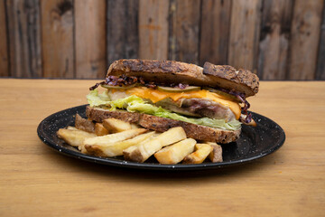 Sandwich. Closeup view of a bun with grilled chicken, lettuce, sliced cabbage, cucumber, cheddar cheese and fries, in a black dish on the wooden table.