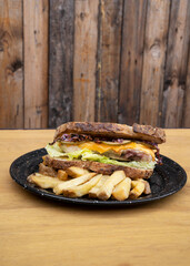 Sandwich. Closeup view of a bun with grilled chicken, lettuce, sliced cabbage, cucumber, cheddar cheese and fries, in a black dish on the wooden table.