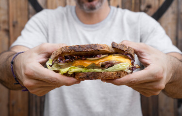 View of a man holding a sandwich made with bread, grilled chicken, lettuce, sliced cabbage, cucumber and cheddar cheese.