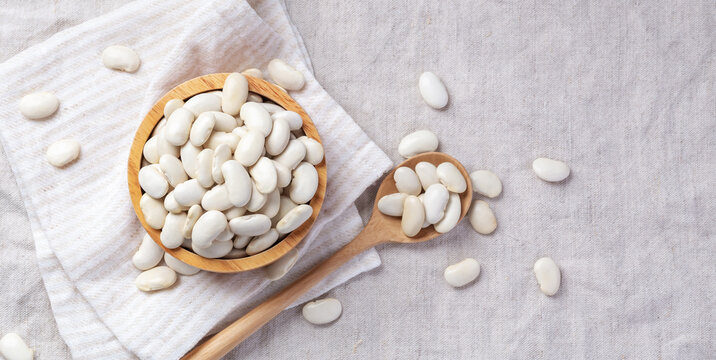 White beans in wooden bowl with spoon on linen tablecloth background, top view