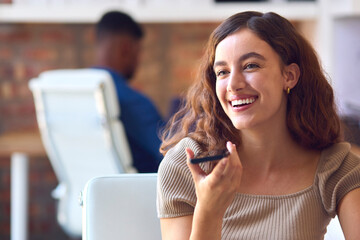 Young Smiling Businesswoman Working At Desk In Office Talking Into Mic Of Mobile Phone
