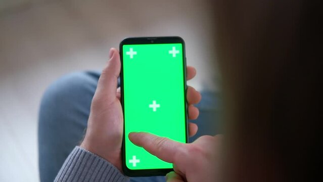 Young man holding smartphone green mock-up screen in hand. Male person using chroma key mobile phone. Vertical mode. Touching, swiping display, tapping, surfing Internet social media. Gesturing