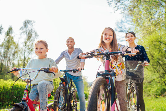 Smiling father and mother with two daughters during summer outdoor bicycle riding. They enjoy togetherness in the summer city park. Happy parenthood and childhood or active sport life concept image.