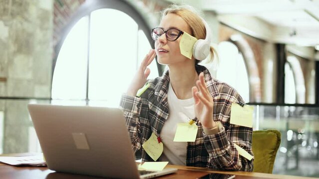 Portrait of cheerful blond woman freelancer with stickers on shirt listen to music in headphones ignore deadline at distance remote work on laptop computer in coworking workplace No stress concept