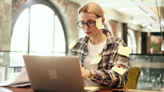 Nervous stressed young blond woman freelancer with stickers typing quickly on laptop computer and looking at paper documents asking to hurry up scream and warning about deadline at coworking workplace