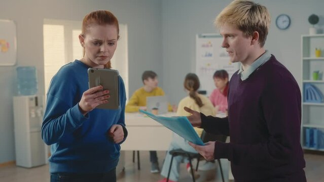 Young man and woman are discussing a new business project, angry and quarreling, comparing data in a digital tablet and a document. Group of young managers working together in a modern office.