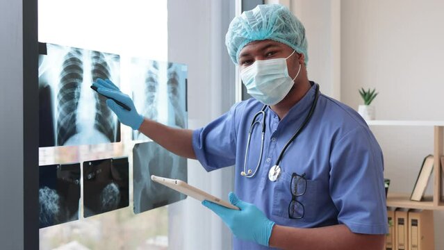 Attentive multiethnic man in medical scrubs and face mask looking at X-ray pictures while holding tablet in doctor's workplace. Physician in gloves and cap examining chest scans in hospital interior.