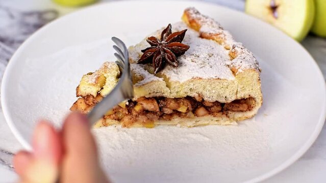 Piece of cinnamon apple pie sprinkled with powdered sugar and decorated with anise star on white plate. Hand breaks off piece of cake with a fork close-up. Delicious dessert in a cafe on a sunny day.