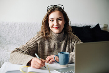 Home office of a young female English tutor with laptop, coffee, and notes, preparing for online lesson with student, sitting at kitchen table. Educator working remotely from home.