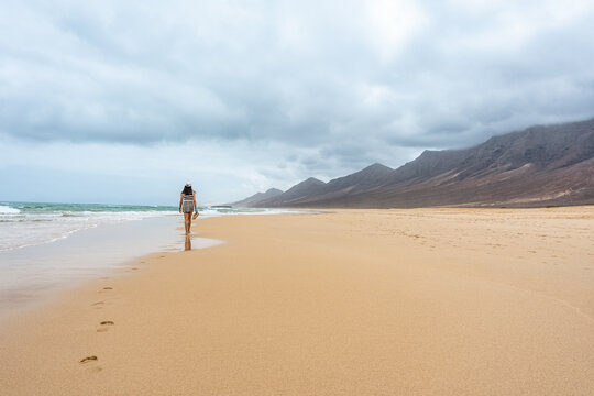 Mujer caminando en playa solitaria de cielo nublado con  chanclas en las manos