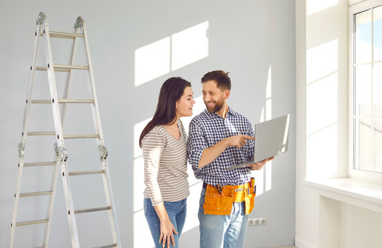 Repairman with laptop talking to young woman. Worker with tool belt standing in room with step ladder, holding modern laptop computer and discussing repairs inside the house with young woman homeowner