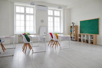 Modern interior of school class without people. Comfortable and ergonomic chairs and desks. Large green chalk board, shelves with accessories for educational process. Clock on the wall.