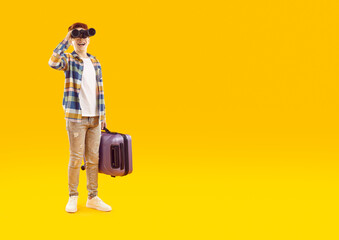 Happy teenage boy with suitcase looking through binoculars. Cheerful casually dressed boy boy holding modern suitcase over isolated copy space studio background. Happy child dreaming about adventures