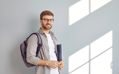 Portrait of smiling male adult student with backpack and folders over grey wall background. Bearded young man in casual clothes, glasses looking at camera. University, college education concept.