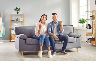 Excited couple raising fists in excitement while sitting on sofa at home. Overjoyed happy young man and woman watching soccer or hockey match on TV, celebrating victory of their team