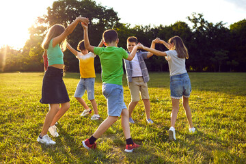 Group of school children playing games and having fun outdoors on a beautiful sunny summer day. Happy friends enjoying good weather, holding hands and dancing a round dance on green grass in the park