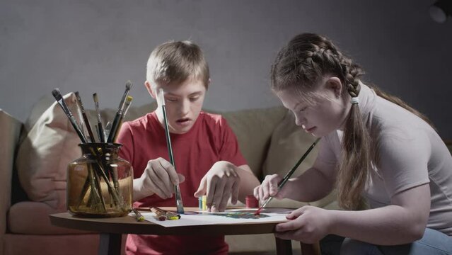 Two children with Down syndrome. A boy and a girl draw with paints and a brush at home. Communication of kids with disabilities. A person with special needs. Chromosomal genetic disorder in a child.