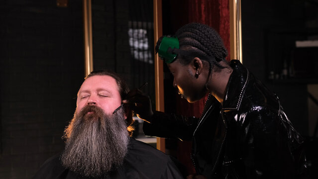 An African-American woman in a black raincoat shaves a man's beard with a straight razor in a barber shop - Powered by Adobe